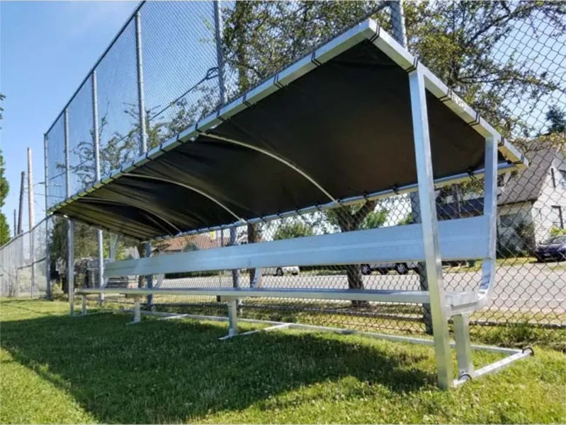Low front angled view of a 21ft aluminum covered bench showing the dark tarp underside beside a chain-link fence.