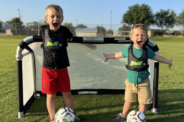Two young kids smiling and posing with soccer balls in front of a portable Quick Fold Aluminum Soccer Goal on a sunny field