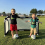Two young kids smiling and posing with soccer balls in front of a portable Quick Fold Aluminum Soccer Goal on a sunny field
