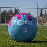 Blue and pink Drip Soccer Ball on turf with a soccer goal blurred in the background