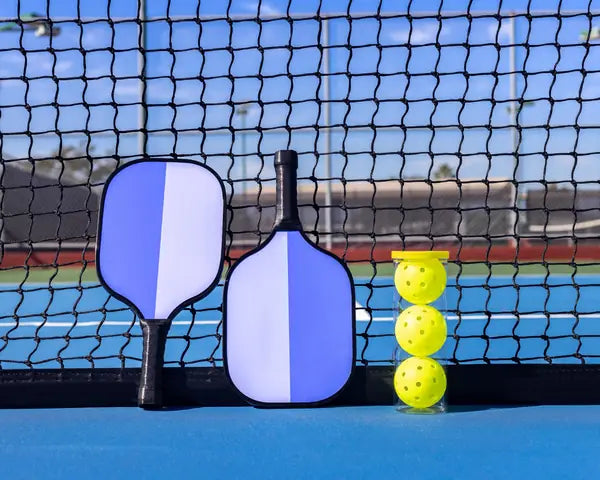 Two blue and white pickleball paddles with three yellow pickleballs displayed in front of a pickleball court net.