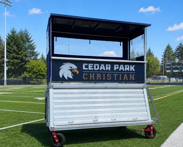 Portable elevated sports press box with a black roof and Cedar Park Christian graphics positioned beside an athletic field.