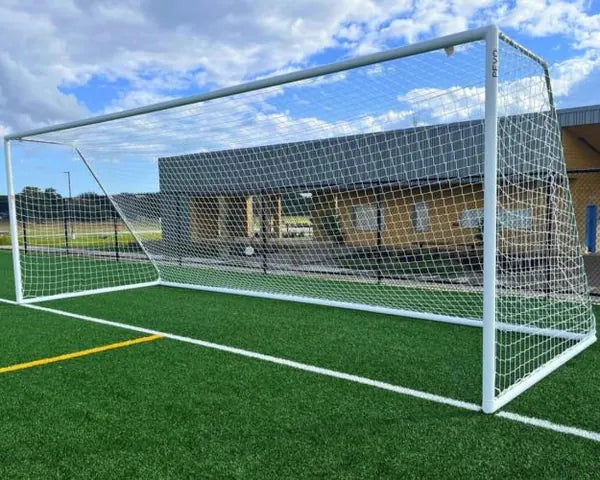 Full-size white aluminum soccer goal with net installed on an artificial turf field beside a school building.