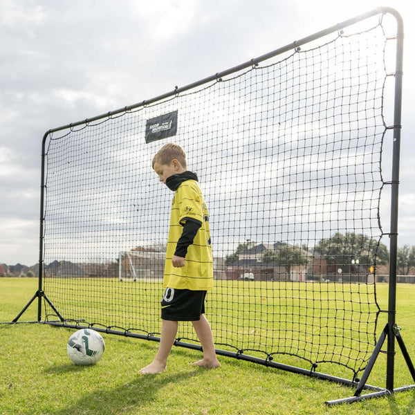 Child in a yellow sports outfit standing next to a large soccer net on a grass field.