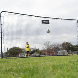 Person standing behind a large black net on a grassy field with trees and buildings in the background.
