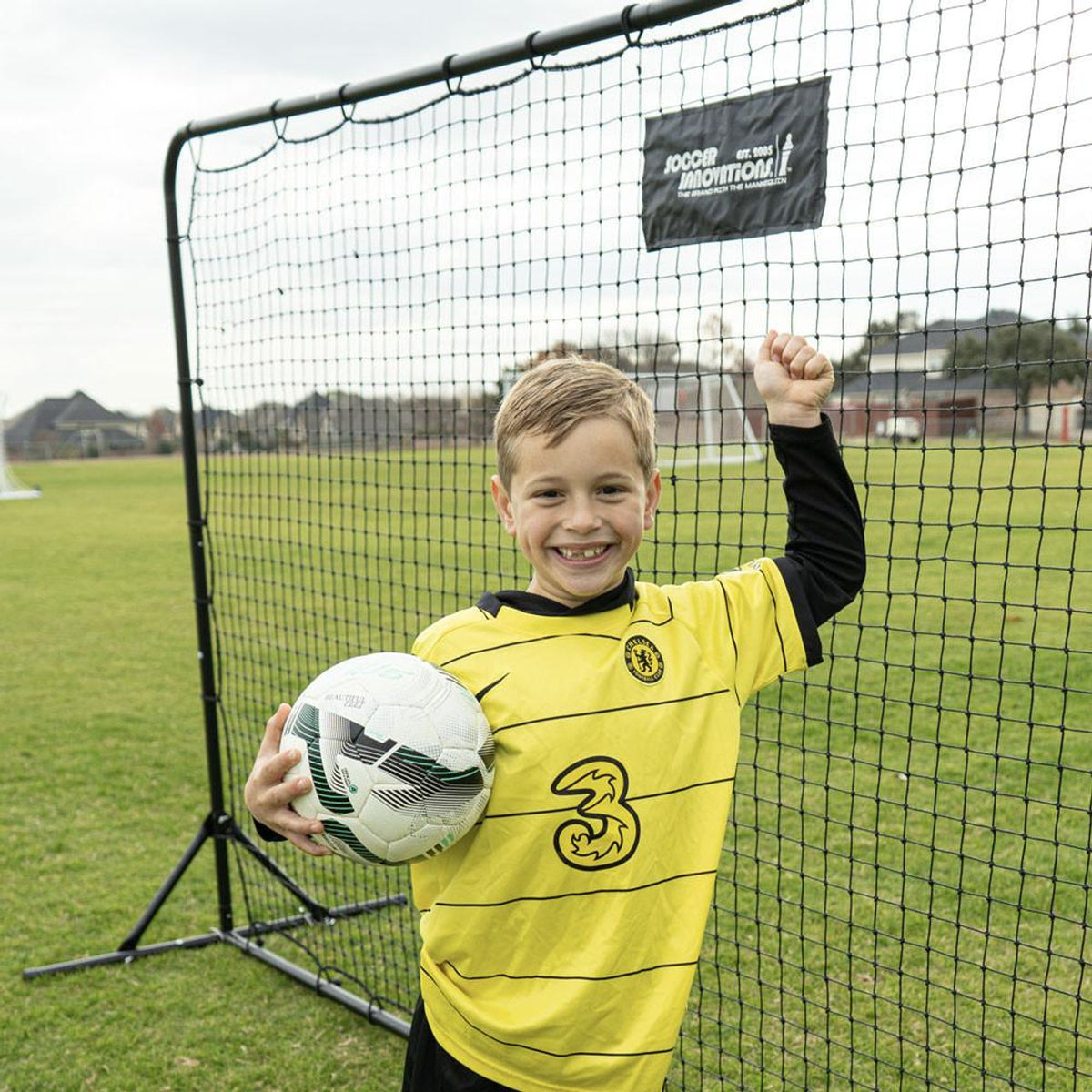 Young boy in a yellow sports jersey holding a soccer ball in front of a net on a grass field.