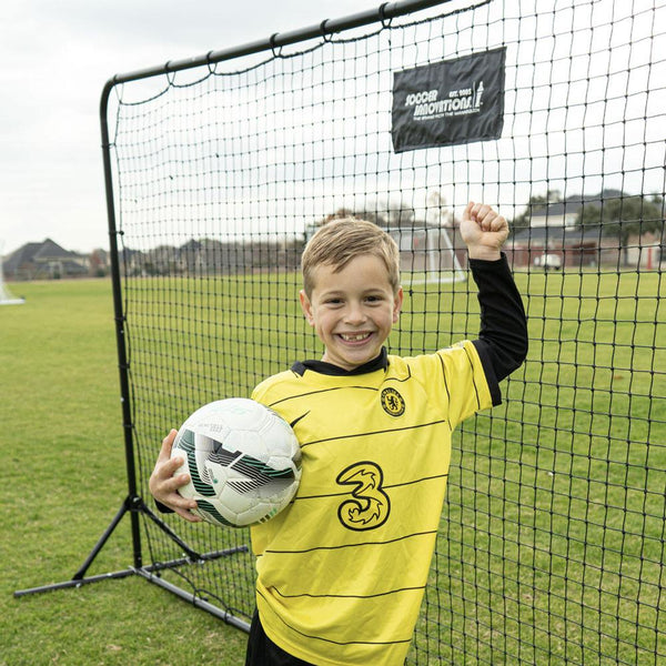 Young boy in a yellow sports jersey holding a soccer ball in front of a net on a grass field.