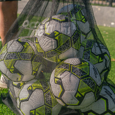 Close-up of black mesh soccer ball bag holding multiple white and neon yellow soccer balls on turf