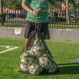 Person carrying a black mesh soccer ball bag filled with soccer balls using the drawstring closure on a turf field