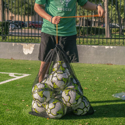 Person carrying a black mesh soccer ball bag filled with soccer balls using the drawstring closure on a turf field