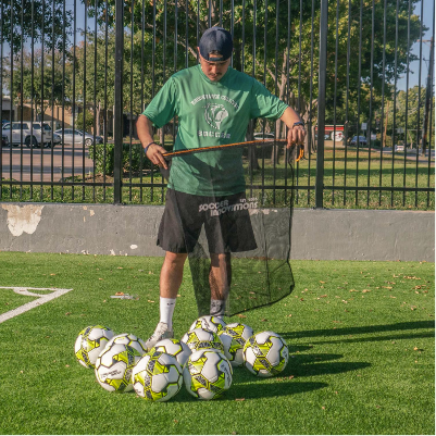 Person opening a black mesh soccer ball bag next to several soccer balls on an outdoor turf field