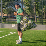 Side view of person carrying a large black mesh soccer ball bag filled with multiple soccer balls on an outdoor field