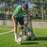 Person loading multiple soccer balls into a black mesh ball bag on artificial turf near a sideline