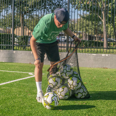 Person loading multiple soccer balls into a black mesh ball bag on artificial turf near a sideline