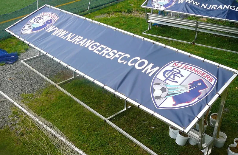 Elevated angled view of an NJ Rangers covered bench with a dark blue tarp, club crest, and white website lettering.